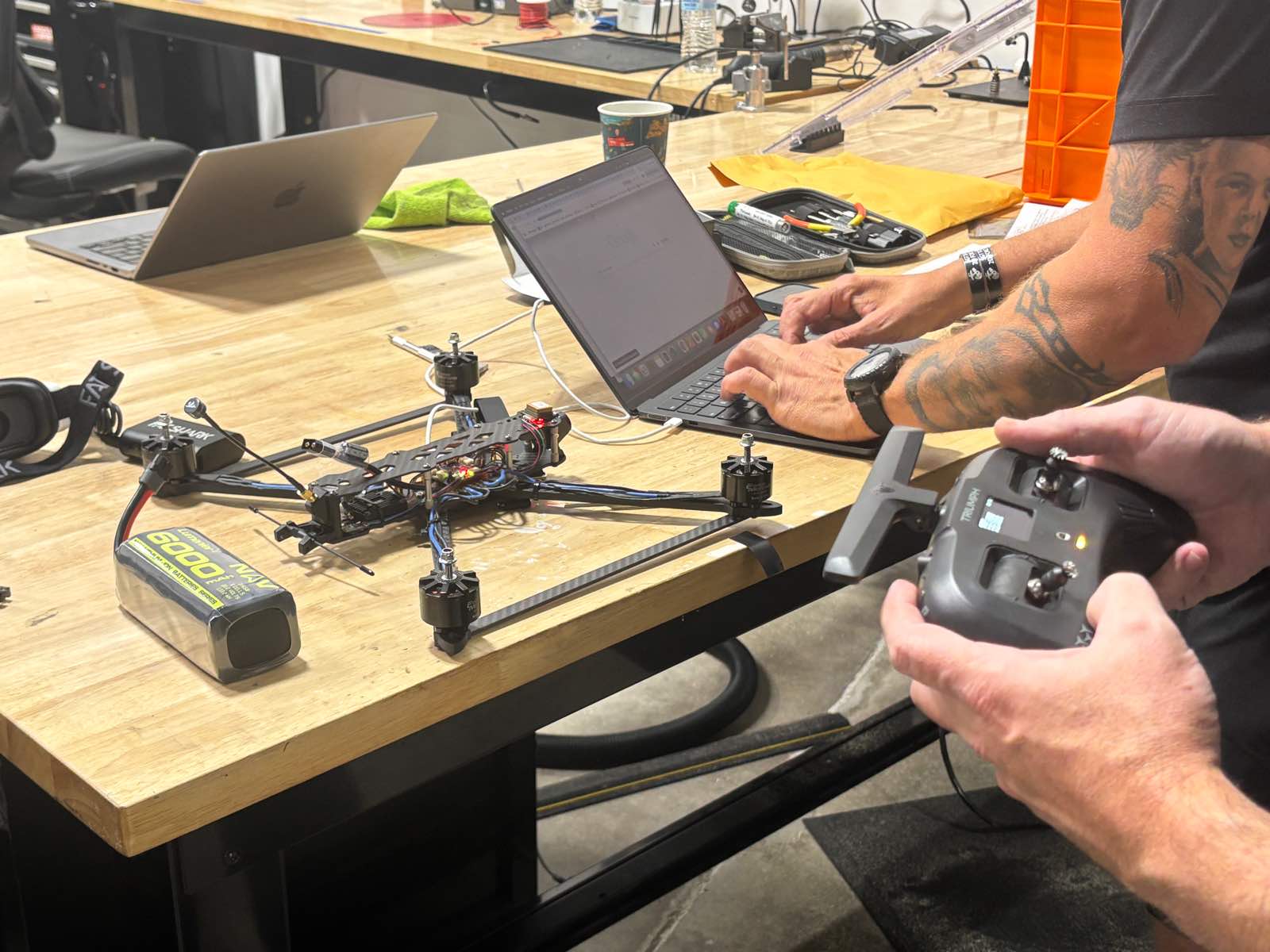 An FPV quadcopter opened up on a workbench next to a laptop, with a battery beside it and a pair of hands holding a transmitter