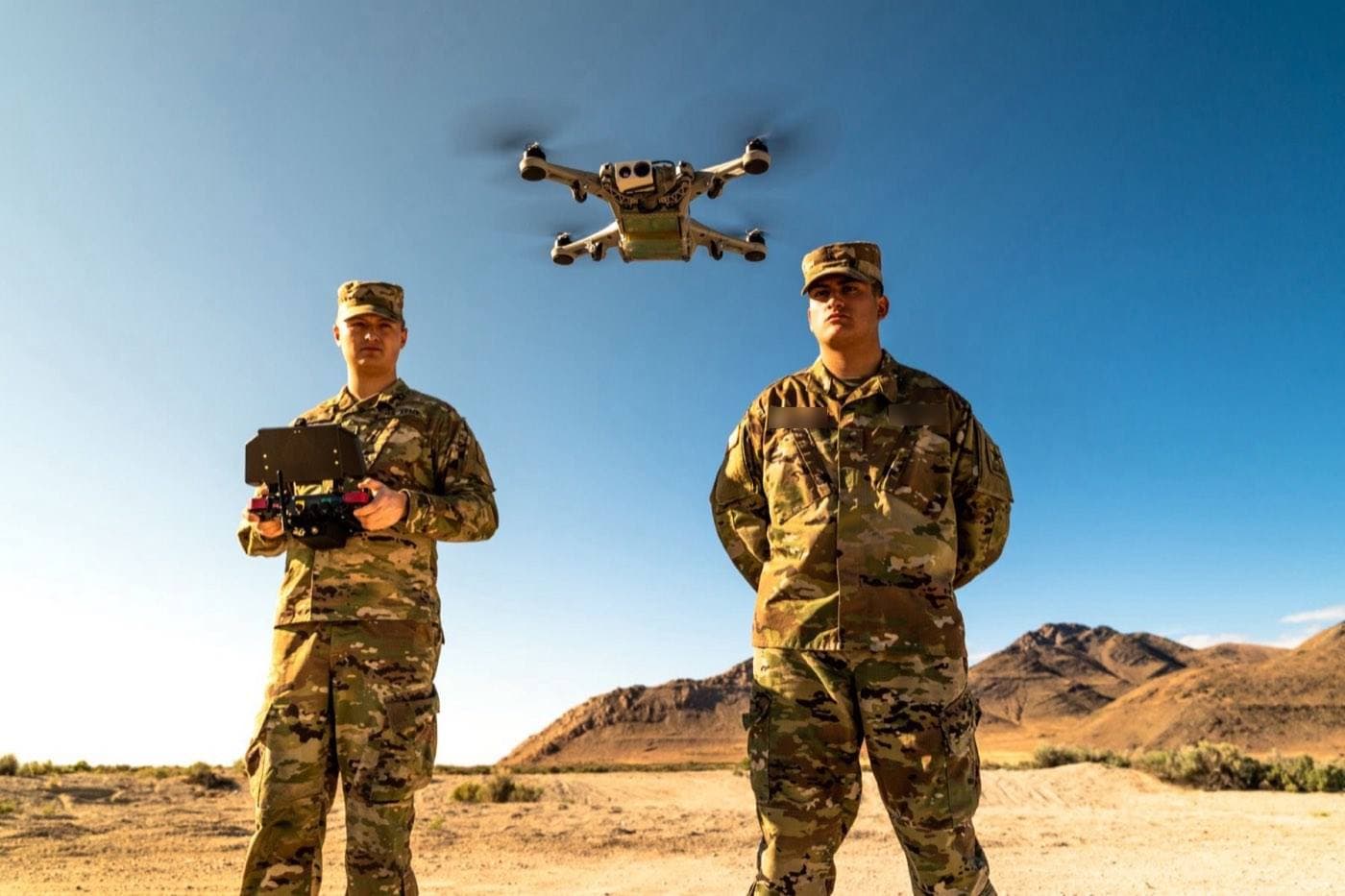 Two operators in uniform in open desert, one at the controls of a small quadcopter hovering above them, name tapes obscured